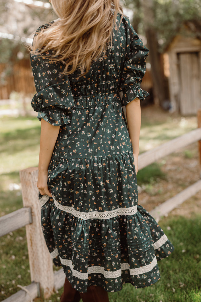A woman with long hair is standing outdoors by a wooden fence, holding the skirt of her Ivy City Co Palermo 3/4 Sleeves Dress in Evergreen Lovelorn Ditsy. The floral cotton dress features lace trim and 3/4 sleeves, creating a graceful and maternity-friendly look amid trees and a wooden shed in the background.