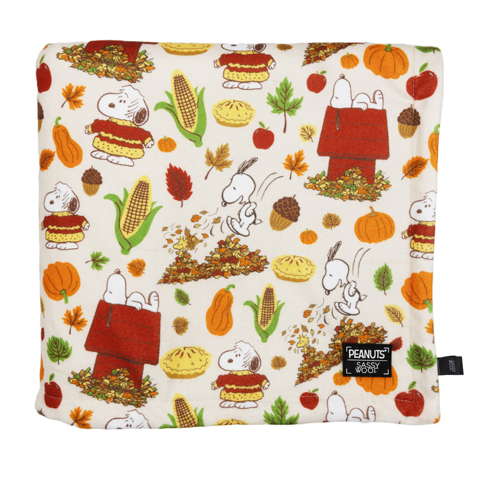 The Sassy Woof Pet Blanket - Peanuts™ Harvest is a soft microfiber throw with autumn-themed Snoopy illustrations—pumpkins, corn, leaves, apples, and acorns—and features a Peanuts x Sassy Woof label in the corner.