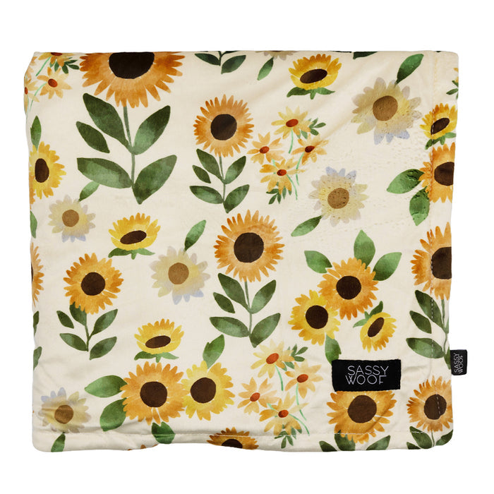 The Pet Blanket - Sunflower Dreams by Sassy Woof is a square, soft polyester dog blanket with a sunflower and green leaf print on cream, finished with a black corner label featuring the Sassy Woof brand name in white.