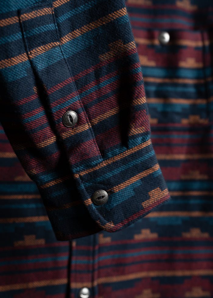 Close-up of a flannel shirt sleeve with a colorful, geometric pattern and metal buttons, highlighting its detailed texture.