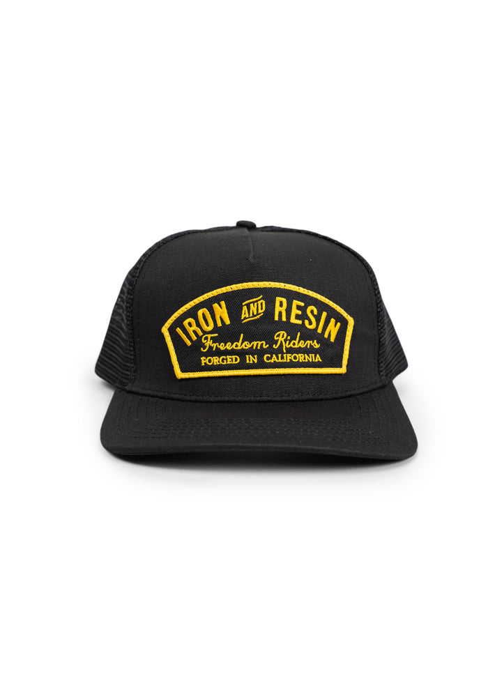 Black trucker hat with yellow "Iron and Resin" patch, reads "Freedom Riders, Forged in California." Simple, bold design.