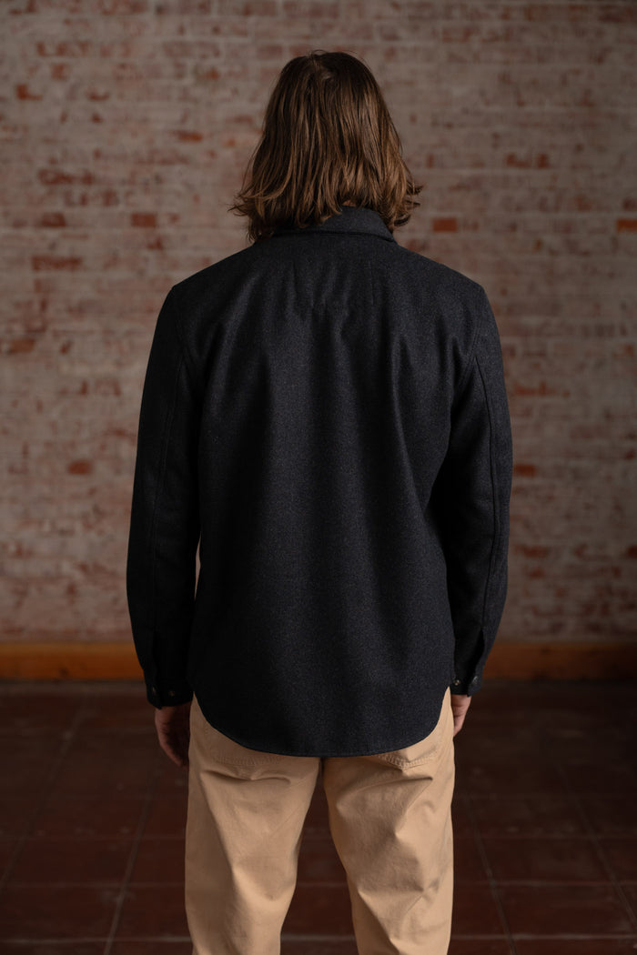 Man with long hair in a dark shirt and beige pants stands facing a brick wall, creating a casual and relaxed atmosphere.