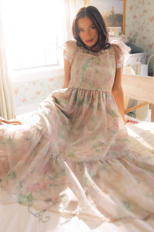A woman wearing the Rhiannon Dress in Painted Bloom by Ivy City Co sits on a bed in a softly lit, vintage-style bedroom with floral wallpaper and sunlight streaming through the window behind her.