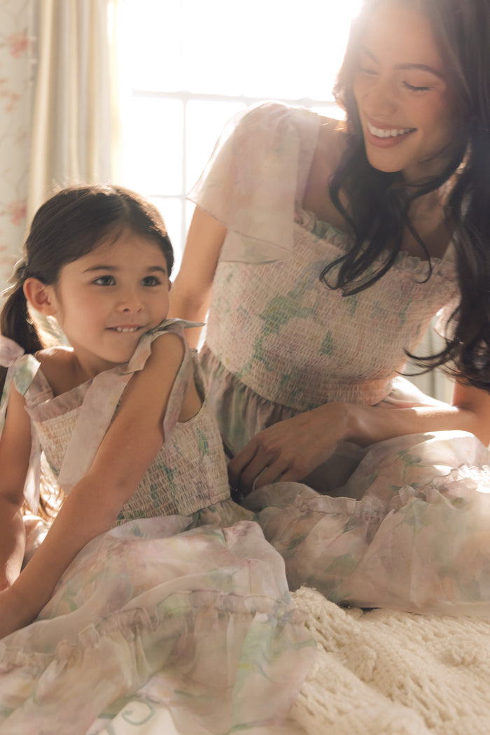 A smiling woman and young girl wearing Ivy City Co’s Rhiannon Dress in Painted Bloom sit together on a bed in soft natural light; the woman gazes warmly at the girl, who looks to the side.