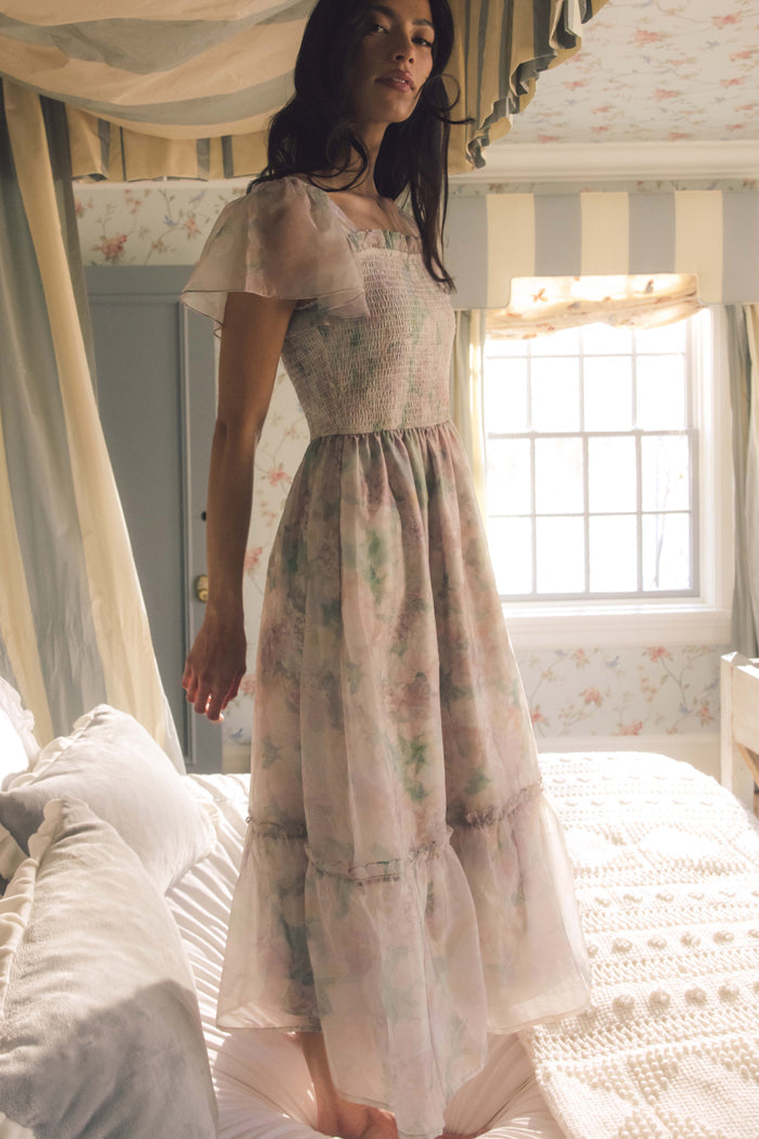A woman in the Ivy City Co Rhiannon Dress in Painted Bloom stands on a bed with a canopy in a softly lit, vintage-style bedroom featuring floral wallpaper.