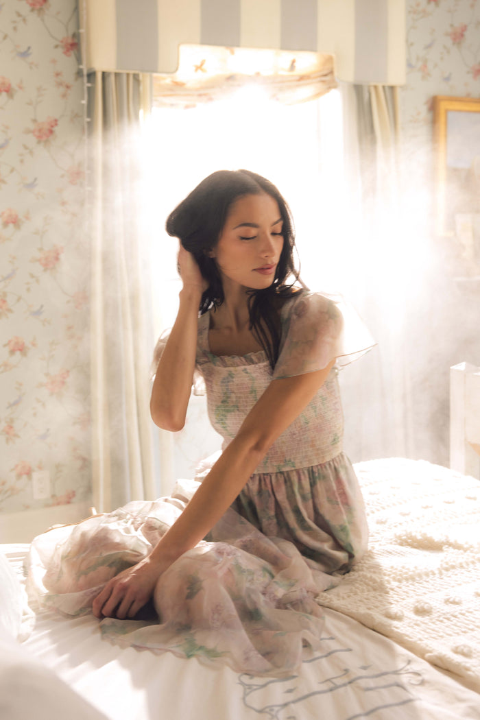 A woman in the Ivy City Co Rhiannon Dress in Painted Bloom sits on a bed, softly lit by sunlight streaming through the window, touching her hair and gazing down thoughtfully.
