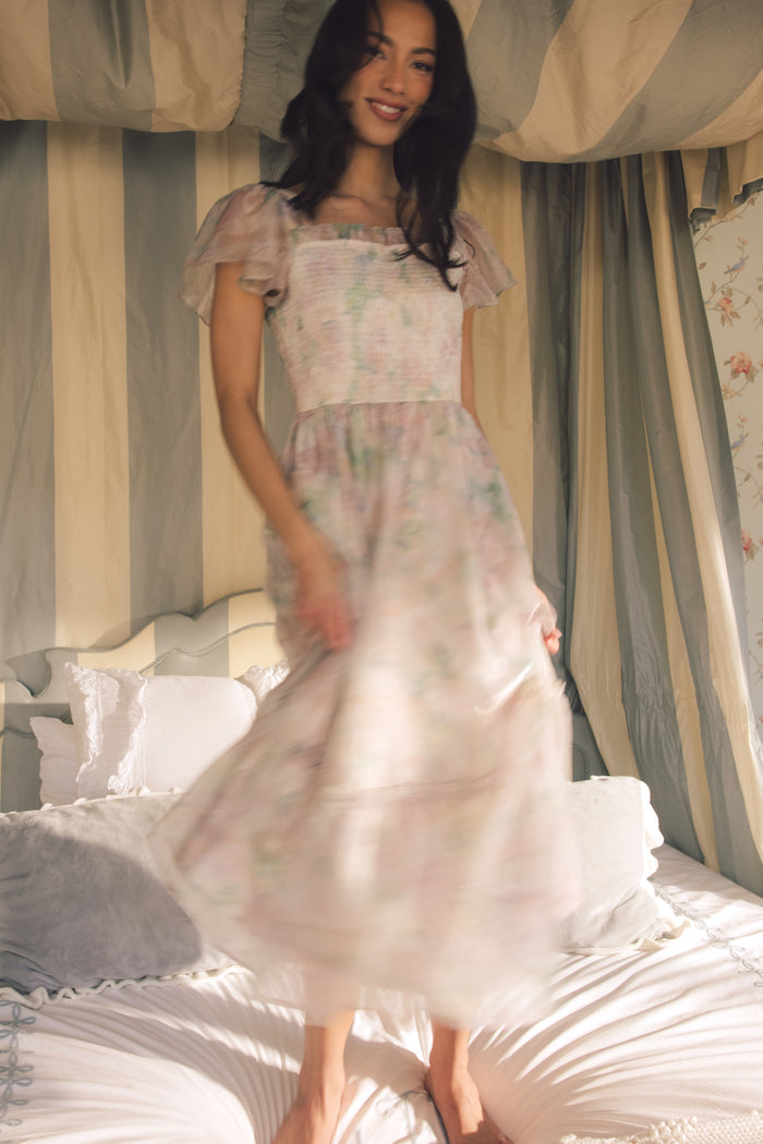 A woman smiles standing on a bed in the Ivy City Co Rhiannon Dress in Painted Bloom—a light, flowy watercolor floral dress. The softly lit room with striped curtains and plush pillows creates a cozy, dreamy atmosphere.