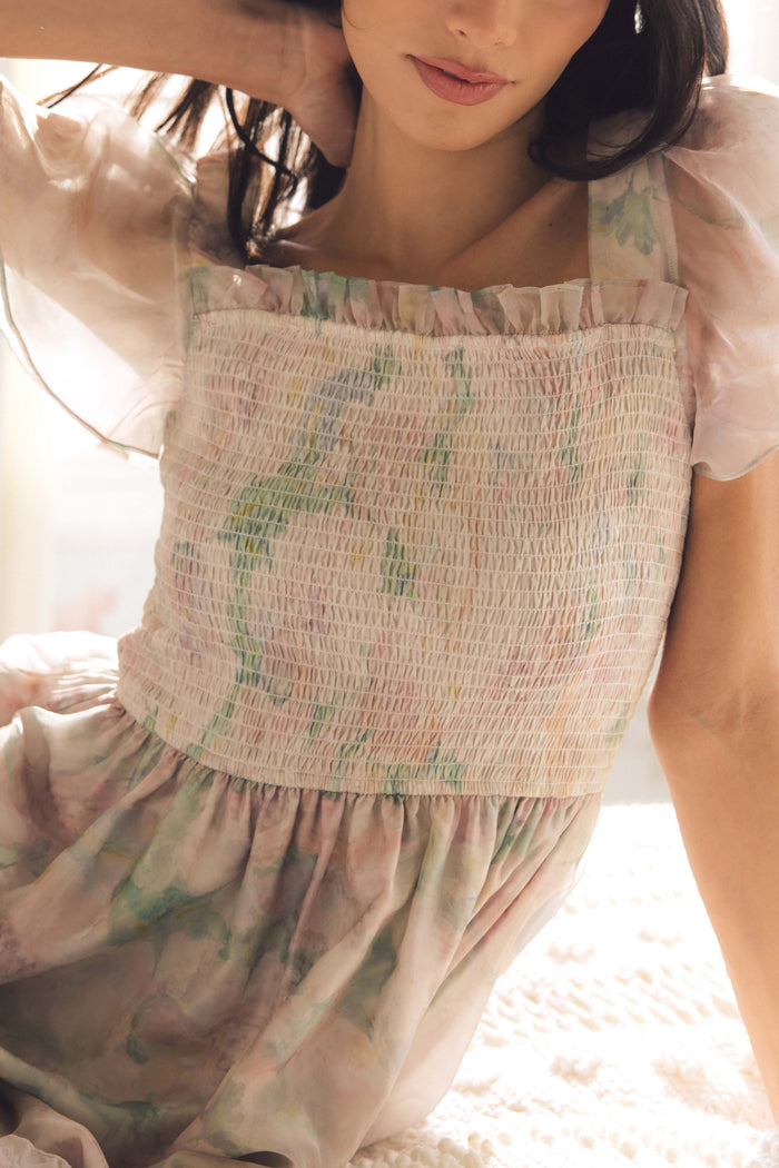A woman in the Rhiannon Dress in Painted Bloom by Ivy City Co, featuring watercolor florals and sheer puff sleeves, sits on a bed. Her dark hair frames her face. This soft, feminine dress is perfect for Easter.