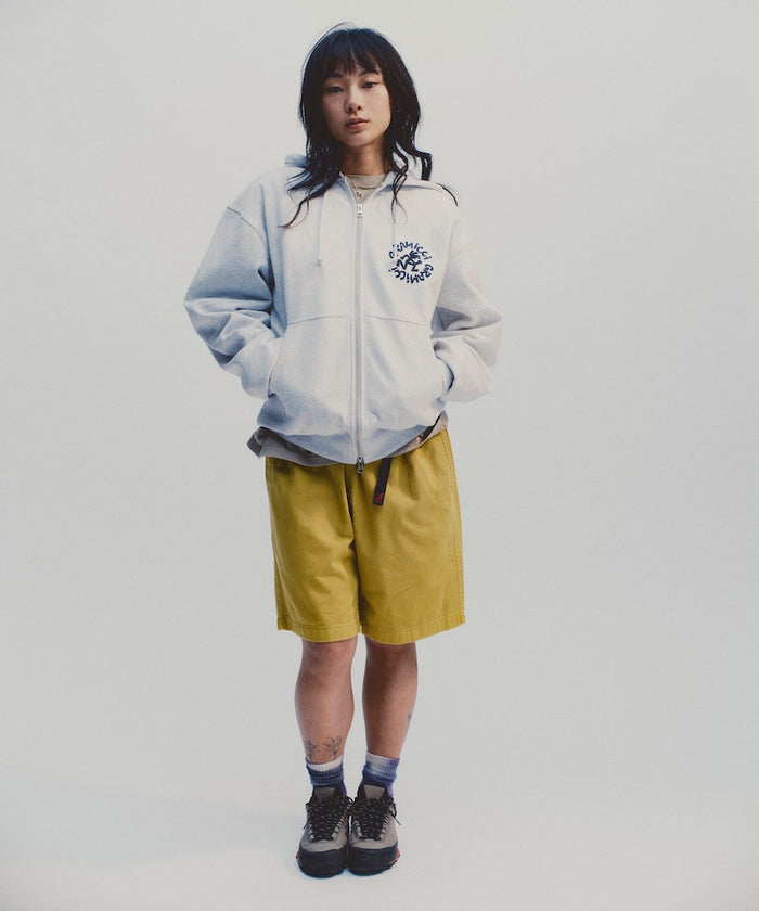 Model wearing heather ash Gramicci Running Man Zip Hooded Sweatshirt and a yellow shorts on a light-tone background.