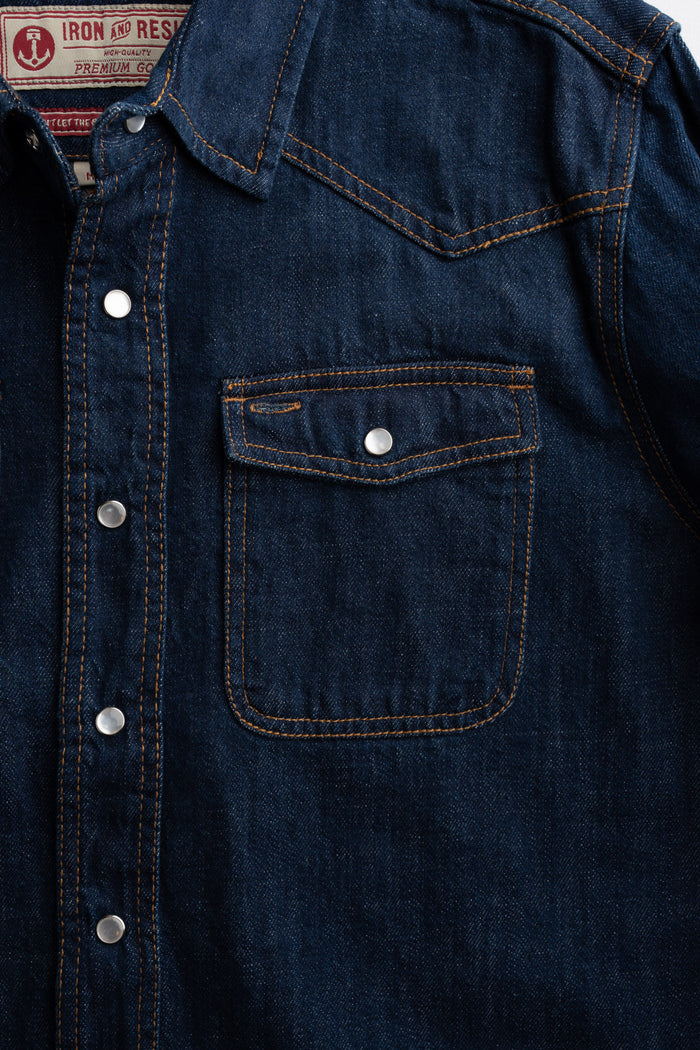 Close-up of a denim shirt with silver buttons and a chest pocket, featuring orange stitching and a visible label inside the collar.