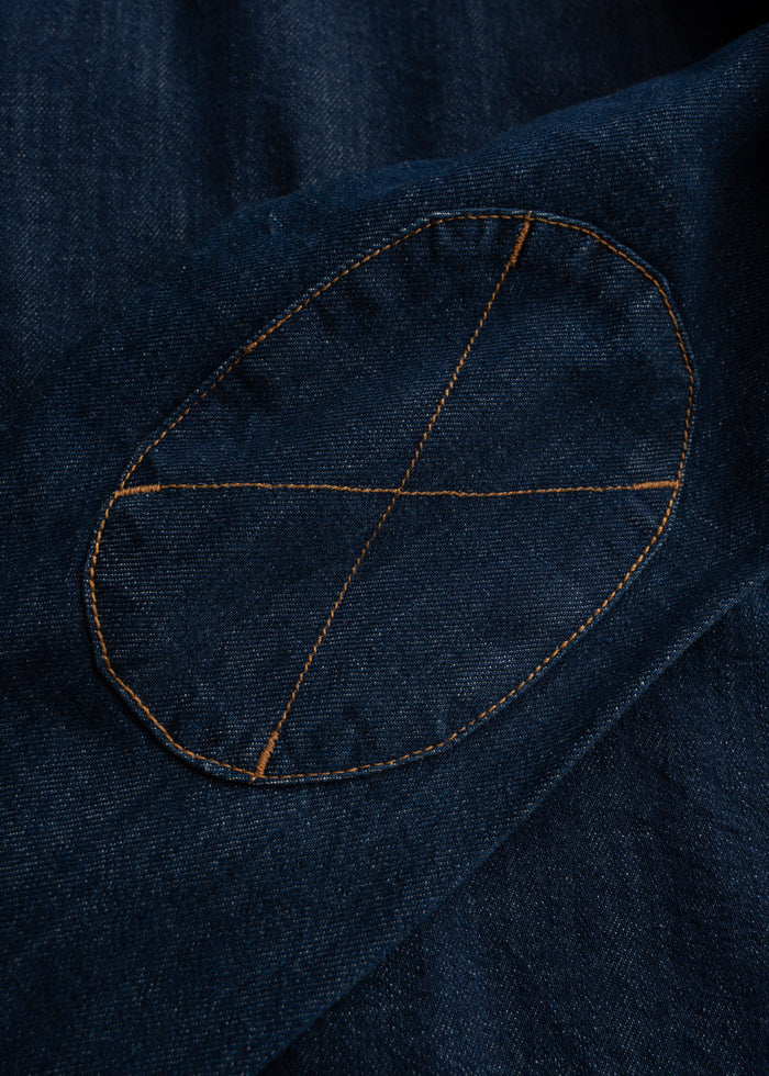 Close-up of a denim jacket's elbow patch with orange stitching, forming an X pattern on dark blue fabric.
