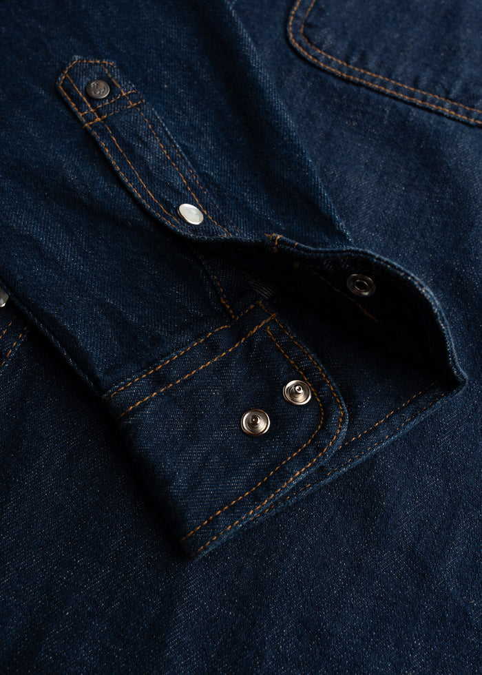 Close-up of a dark denim shirt with visible orange stitching and metal snap buttons, showcasing detailed craftsmanship.