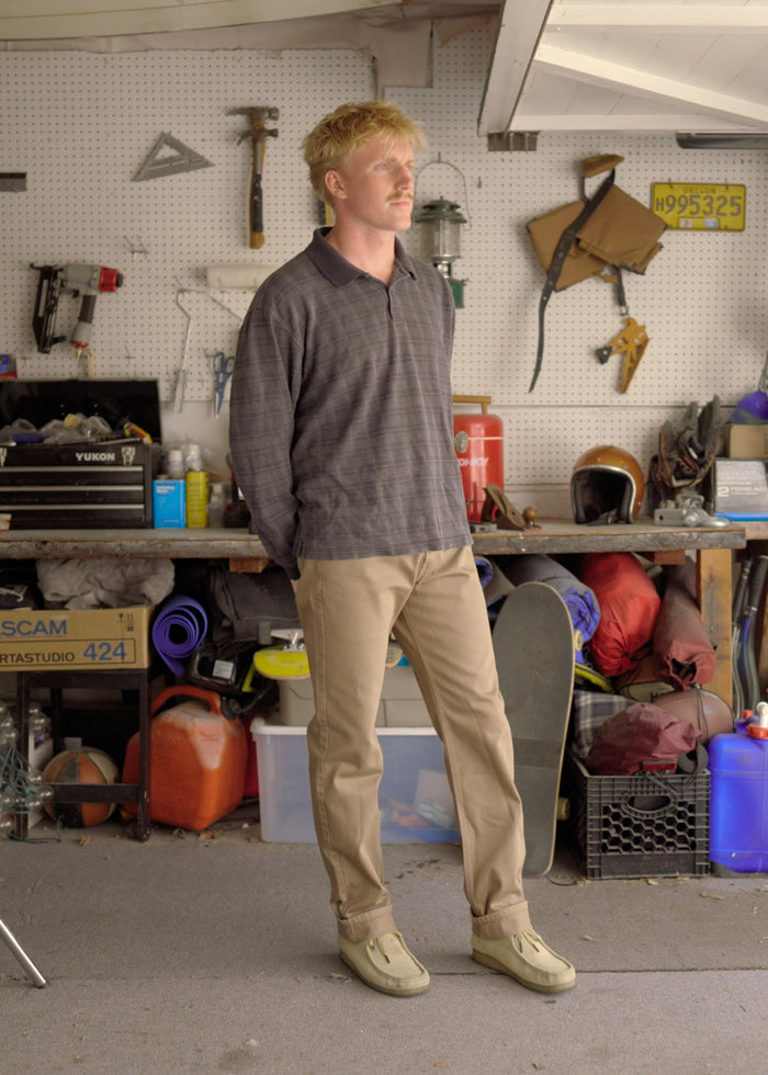 Man in a garage leans against a workbench, surrounded by tools and gear, wearing a plaid shirt and khaki pants, looking thoughtful.