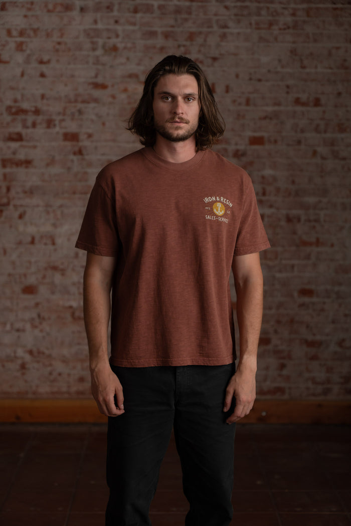 Man in brown "Iron & Resin" t-shirt stands against a brick wall, looking forward. Casual and relaxed atmosphere.