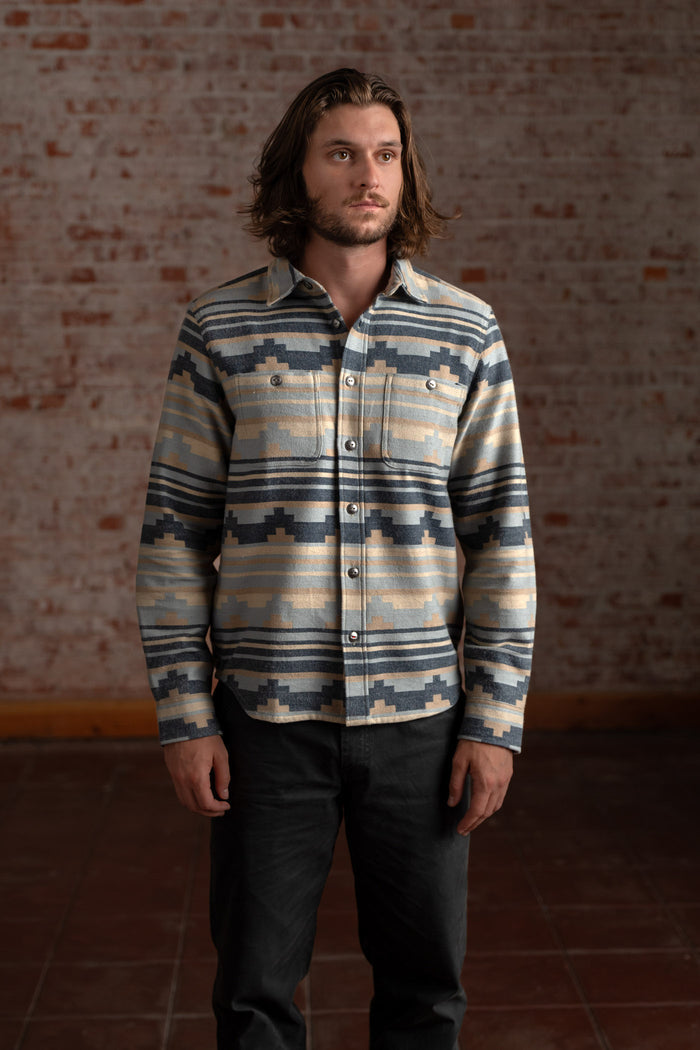 Man with long hair wears a patterned shirt and dark pants, standing against a rustic brick wall in a dimly lit room.
