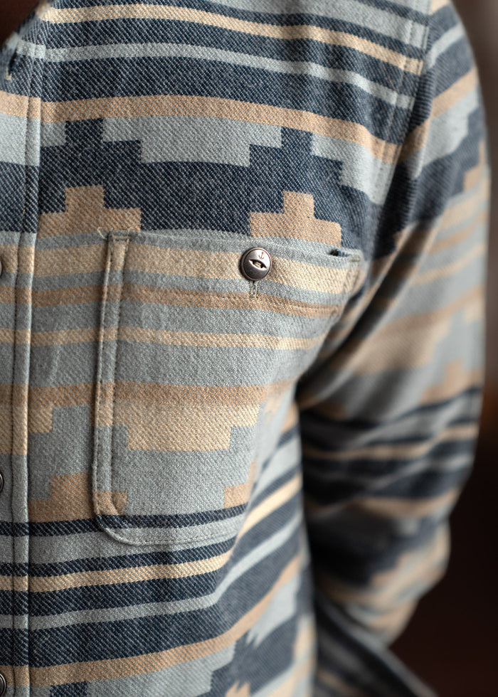 Close-up of a patterned shirt with geometric designs and a buttoned pocket, featuring blue, beige, and gray tones.