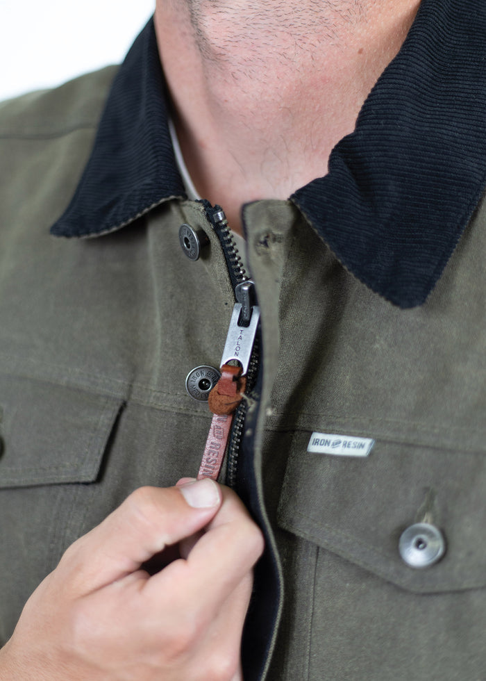 A person zips up a green jacket with a black collar, showcasing an Iron and Resin label and a leather zipper pull.