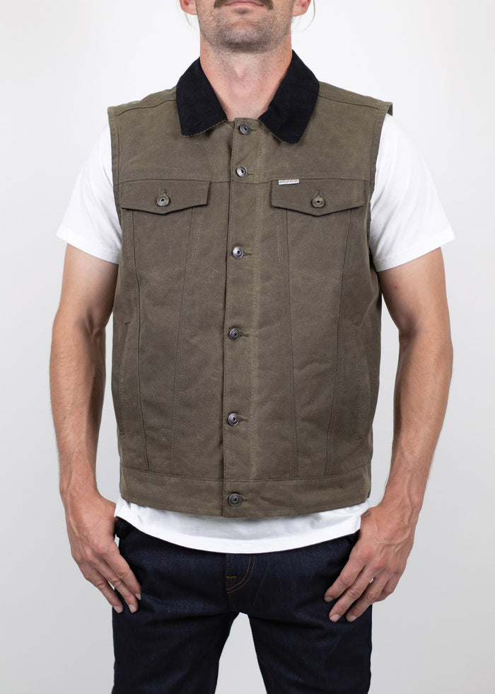 Man in a brown buttoned vest over a white shirt, hands in pockets, standing against a plain background, showcasing casual style.