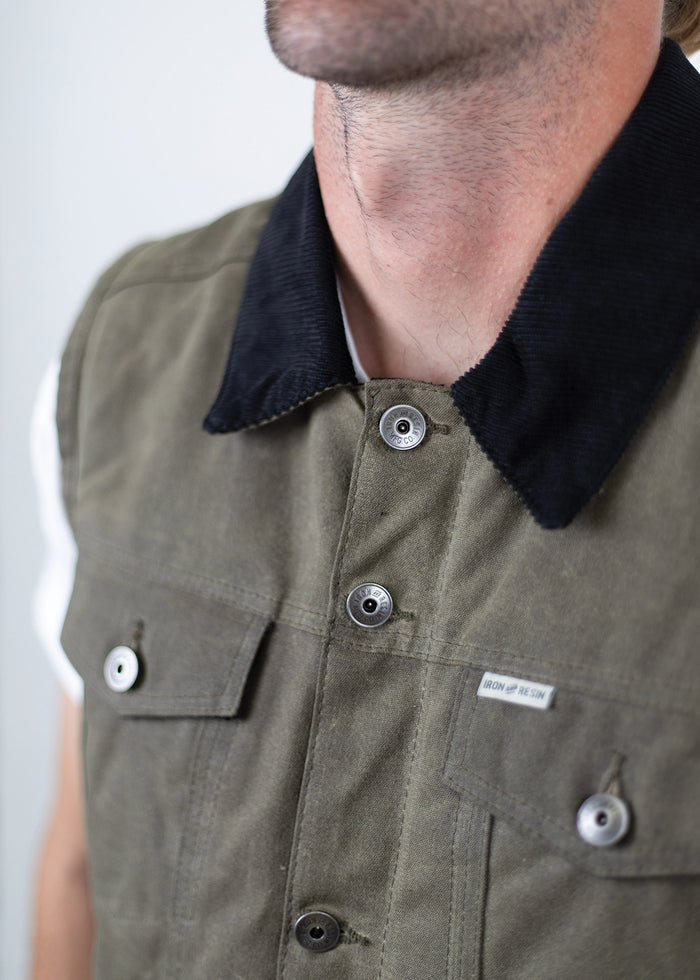 Close-up of a man wearing an olive green vest with a black collar and buttons, featuring a small "Iron and Resin" label.
