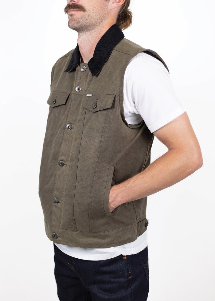 Man in a brown vest with black collar, white shirt, and jeans, standing with hands in pockets against a plain white background.