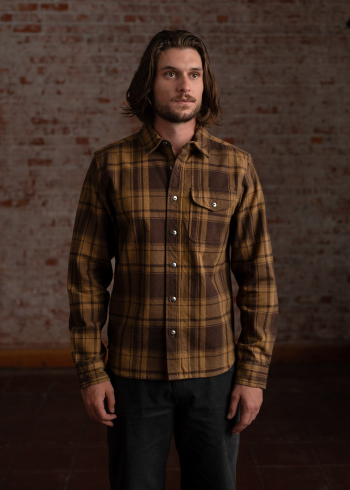 A man in a brown plaid shirt stands against a brick wall, looking to the side with a thoughtful expression.