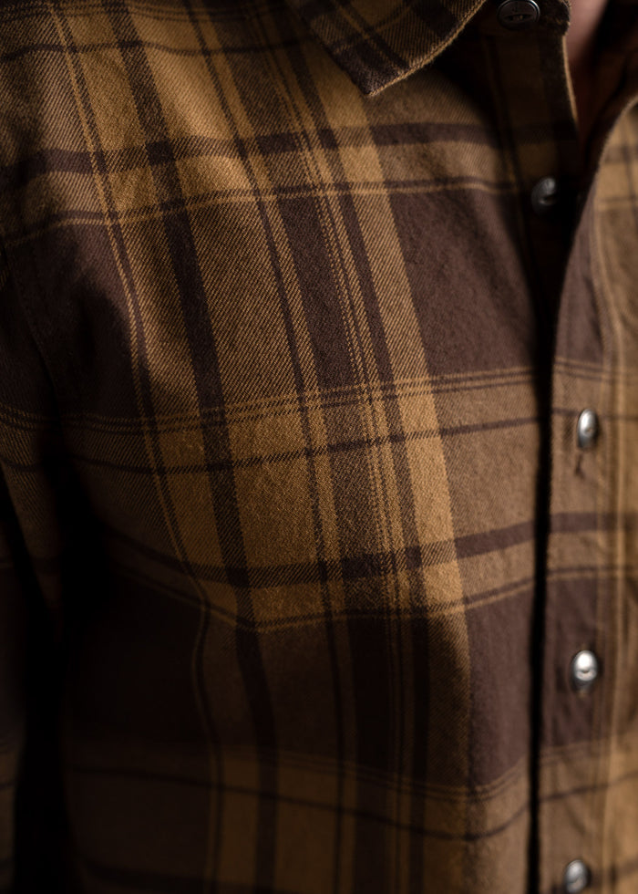 Close-up of a brown plaid shirt with button details, highlighting the fabric texture and pattern in warm lighting.