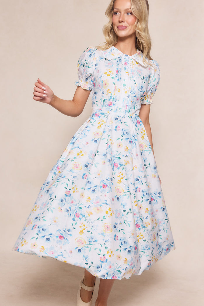 A woman poses and smiles against a neutral background, wearing the Ivy City Co Sophia Dress in Blue Bouquet—a short-sleeve, nursing-friendly dress with a fitted waist and full skirt.