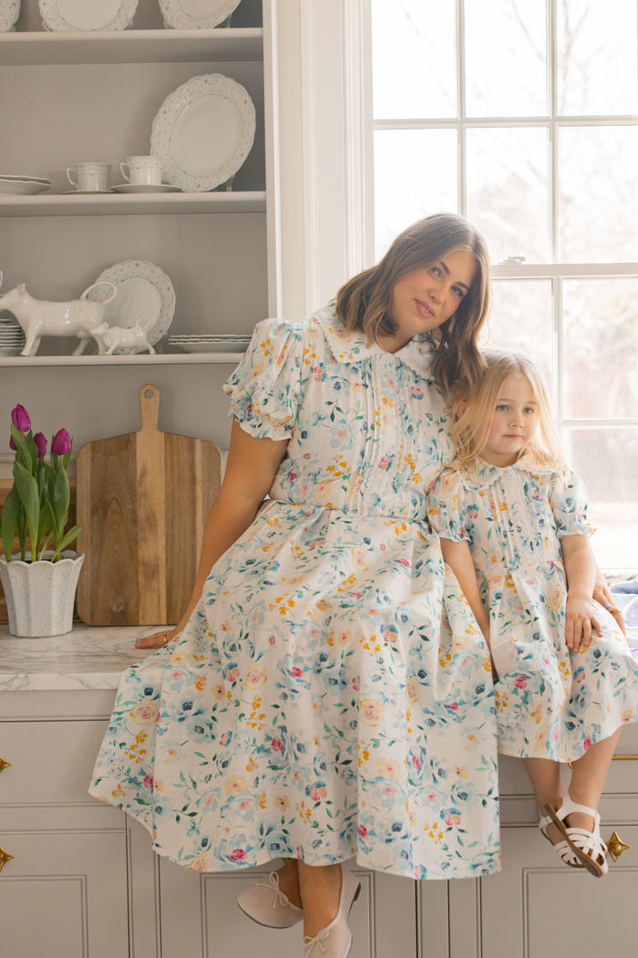 A woman and a young girl wear matching Ivy City Co Sophia Dresses in Blue Bouquet as they sit on a kitchen counter beside purple tulips, with sunlight streaming in and white dishes lining the shelves behind them.