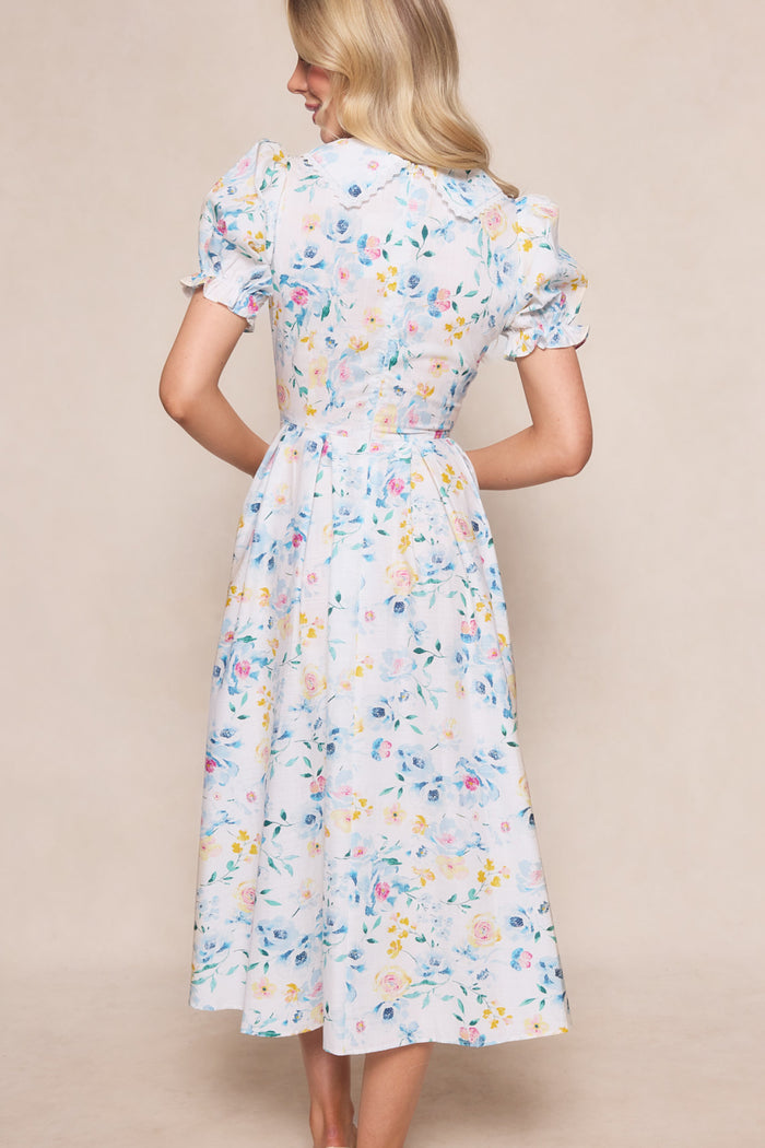 A woman with long blonde hair faces away, wearing the Ivy City Co Sophia Dress in Blue Bouquet—a nursing-friendly, floral midi dress with short puff sleeves and a fitted waist that flows to mid-calf, set against a soft beige background.