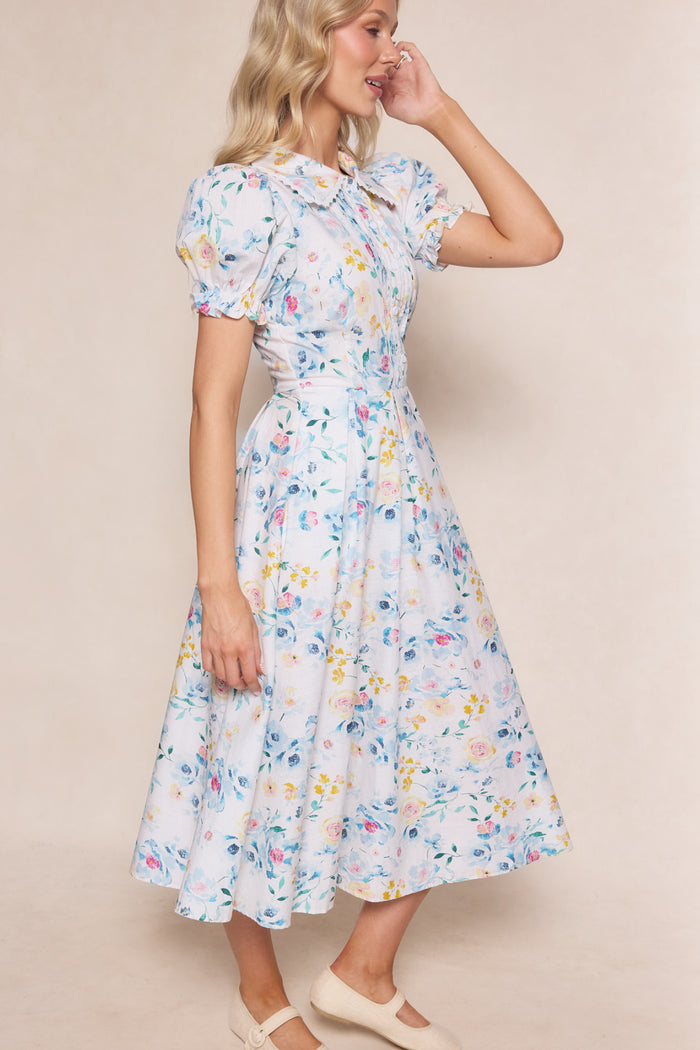 A woman with wavy blonde hair and beige shoes stands sideways, smiling with one hand on her face, wearing the Ivy City Co Sophia Dress in Blue Bouquet—a floral print nursing friendly dress with short puff sleeves and a flared midi skirt.