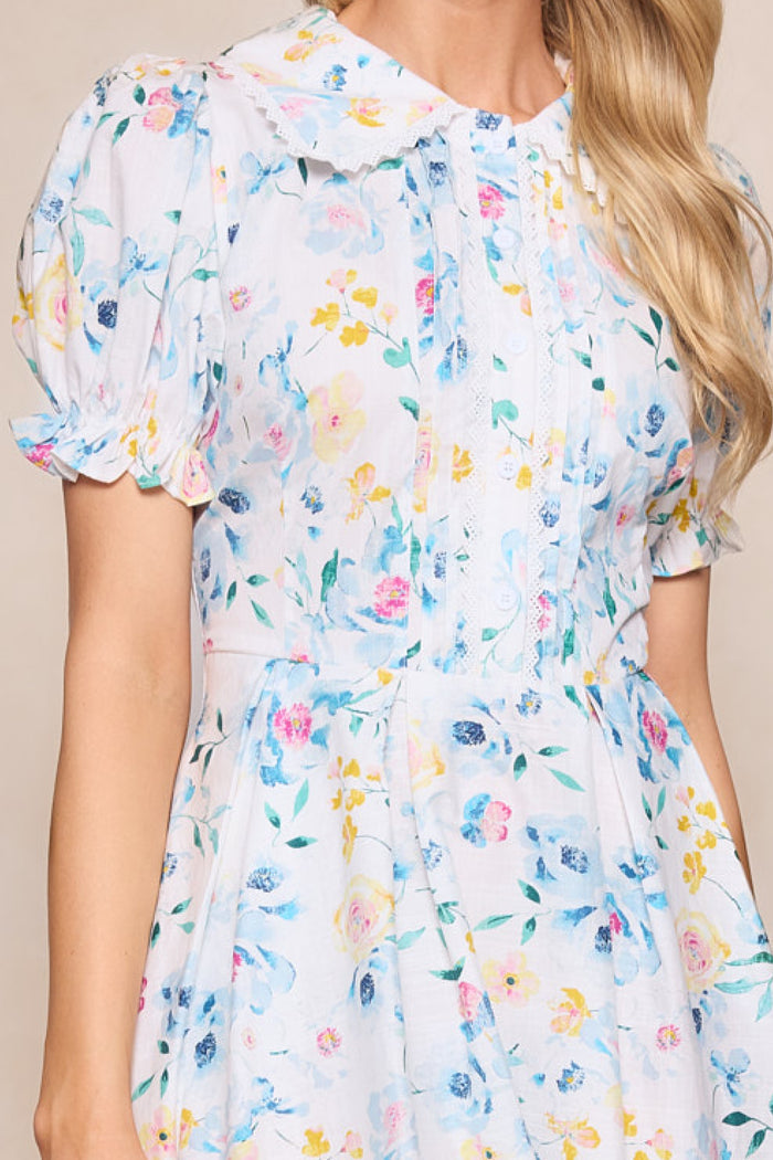 A woman wears the Sophia Dress in Blue Bouquet by Ivy City Co—a charming midi with pastel florals, puffed sleeves, Peter Pan collar, decorative buttons, and lace; perfect for any occasion.