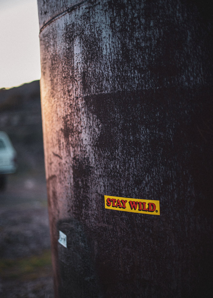 Rusty metal tank with "Stay Wild" sticker in focus, car blurred in background, capturing a rugged outdoor vibe at dusk.