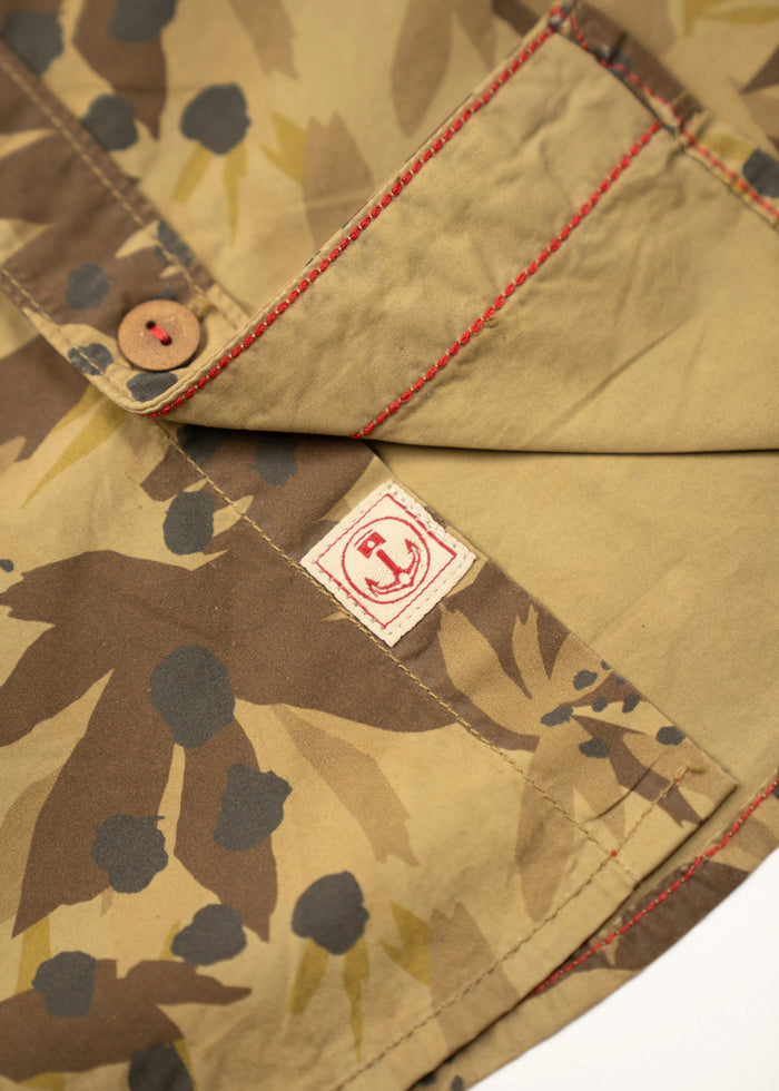 Close-up of a camouflage shirt with red stitching and a button, featuring a small red logo tag on the fabric.