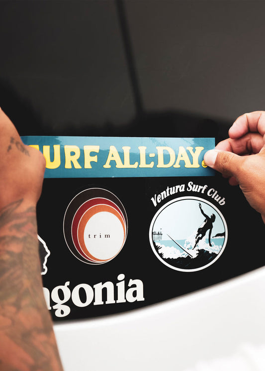 Hands apply "Surf All-Day" sticker above Trim and Ventura Surf Club decals on a black surface.