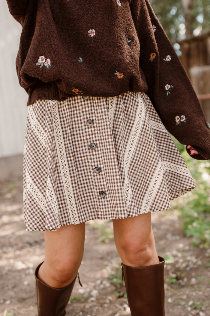 A person stands outdoors on a sunny day wearing a brown floral sweater, the Ivy City Co Tash Skort in Hickory Gingham featuring buttons and lace trim accents, and knee-high brown boots.