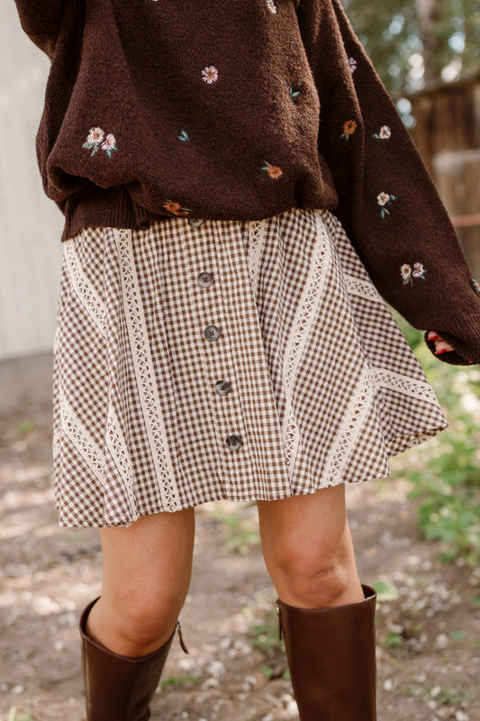 A person stands outdoors on a sunny day wearing a brown floral sweater, the Ivy City Co Tash Skort in Hickory Gingham featuring buttons and lace trim accents, and knee-high brown boots.