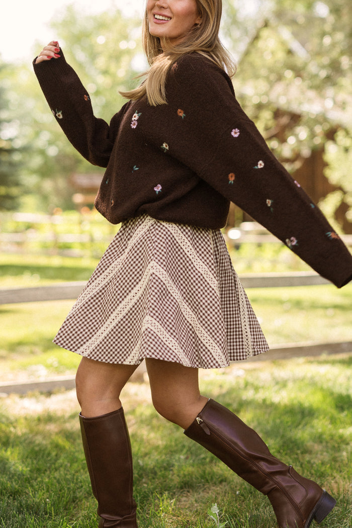 A woman twirls outside on the grass in the Ivy City Co Tash Skort in Hickory Gingham, paired with a brown sweater featuring floral details and tall brown boots. Trees and a wooden fence serve as the backdrop.