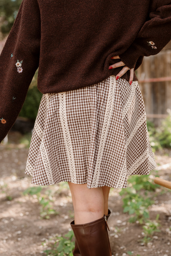 A person stands outdoors, their lower half visible and dressed in tall brown boots, red nail polish, a brown sweater, and the Ivy City Co Tash Skort in Hickory Gingham—a brown checked cotton-blend skort with lace trim details.