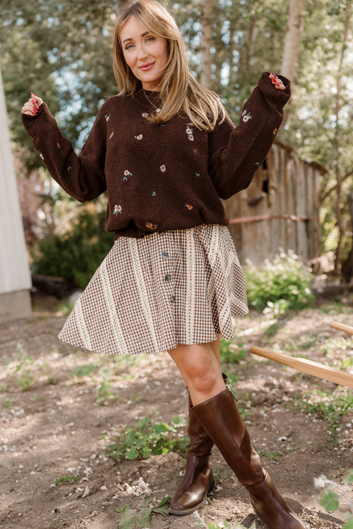 A woman stands outdoors in front of trees and a wooden shed, smiling as she poses with one leg slightly forward. She wears a brown sweater with small floral patterns, knee-high brown boots, and the Ivy City Co Tash Skort in Hickory Gingham—a cotton-blend skort featuring delicate lace trim.