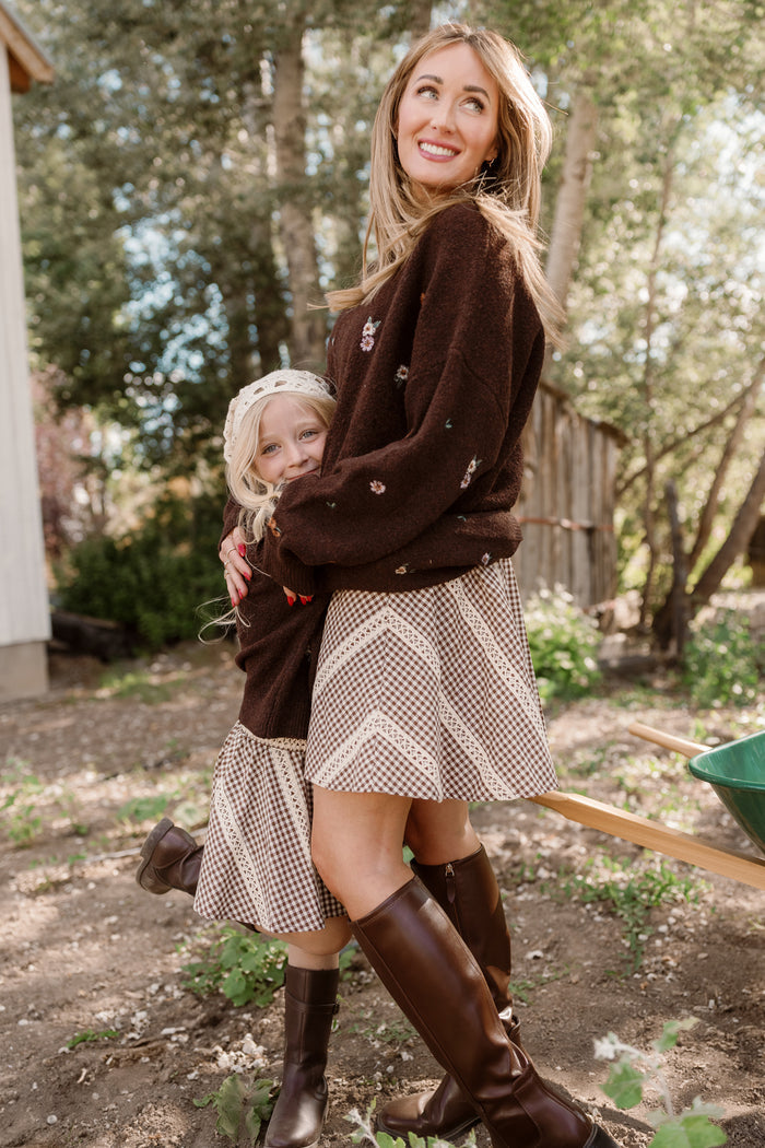 A woman and a young girl, both wearing matching brown sweaters and Ivy City Co’s Tash Skort in Hickory Gingham with delicate lace trim, hug and smile outdoors in a sunlit garden with trees and a wooden shed in the background.