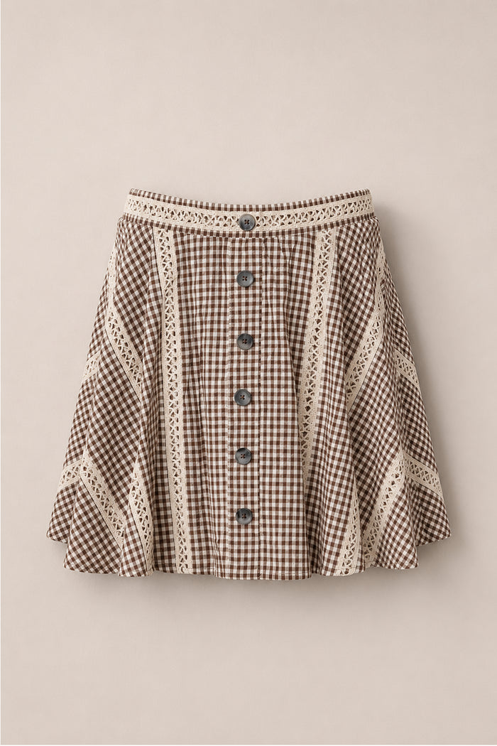 The Tash Skort in Hickory Gingham by Ivy City Co, featuring a brown and white gingham pattern, cream lace trim, front buttons, and an A-line silhouette, is displayed against a light beige background.
