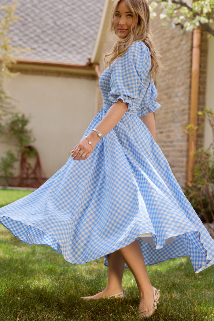 A woman with long, wavy blonde hair twirls outside on the grass, smiling softly. She wears tan flats and the Teagan Dress in Blue Gingham by Ivy City Co. The background features a house and trees. This charming dress is both stylish and maternity friendly, combining comfort with effortless elegance.