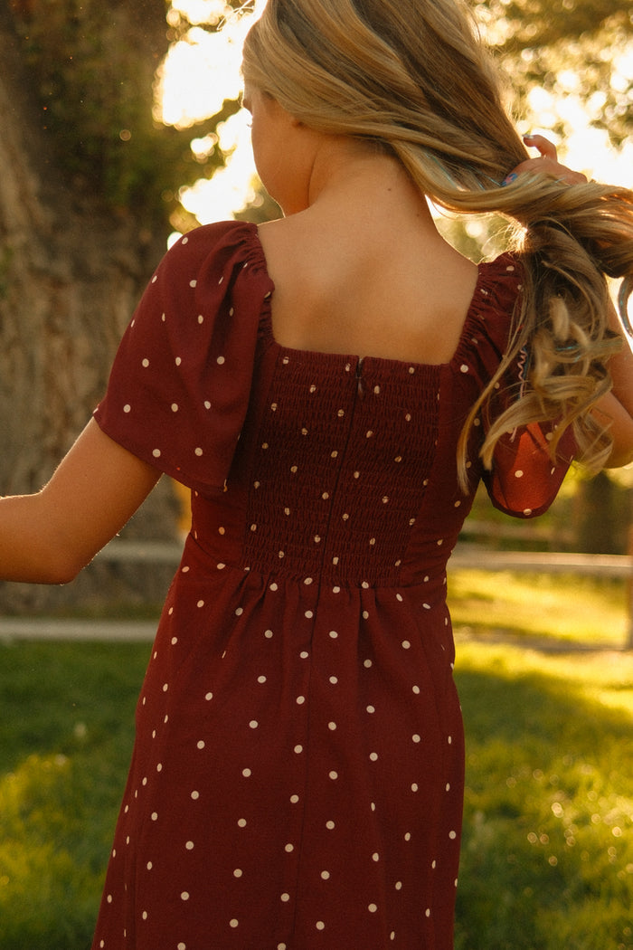 A person with long blonde hair, wearing the Ivy City Co Teen Blakely Dress in Cranberry Dainty Dot—featuring a maroon midi length with white polka dots and delicate petal sleeves—stands outdoors in the sunlight, facing away and gently holding their hair, while green grass and trees provide a scenic background.