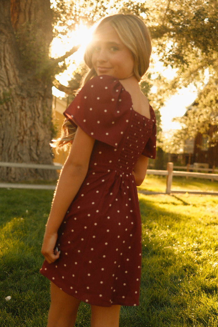 A young woman stands outdoors on the grass, sunlight glowing behind her, wearing the Ivy City Co Teen Blakely Dress in Cranberry Dainty Dot—a charming midi-length style featuring petal sleeves and playful polka dots—as she looks back over her shoulder.