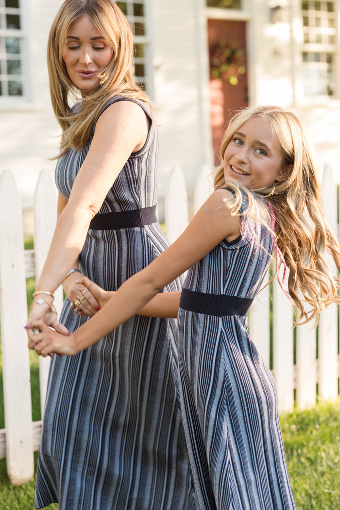 A woman and a young girl smile and hold hands outdoors in front of a white picket fence and house, both wearing matching Ivy City Co Teen Laurel Dresses in Midnight.