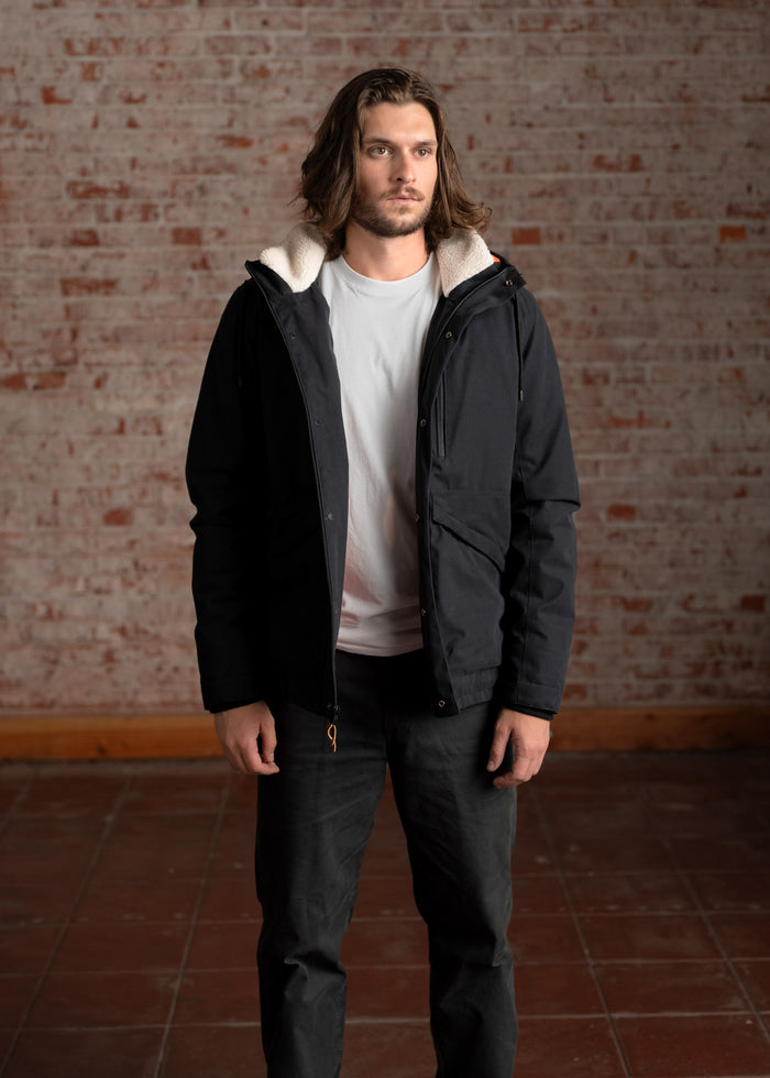 Man in a black jacket and white shirt stands against a brick wall, gazing forward with a thoughtful expression.