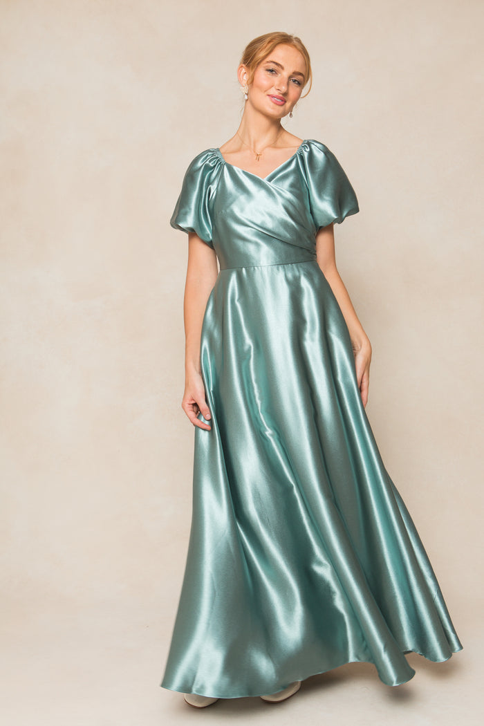 A woman wears the Ivy City Co Tessie Dress in Seaglass Satin, featuring short puff sleeves and a flowing, floor-length skirt—an ideal fall bridesmaid dress—standing against a neutral beige background.