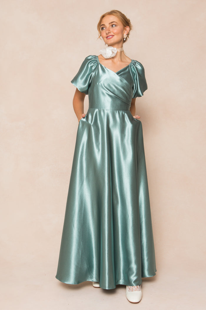 A woman smiles in the Ivy City Co Tessie Dress in Seaglass Satin, featuring short puff sleeves, pockets, and a shiny finish. She accessorizes with white shoes and a white flower choker—an elegant choice for fall bridesmaids.