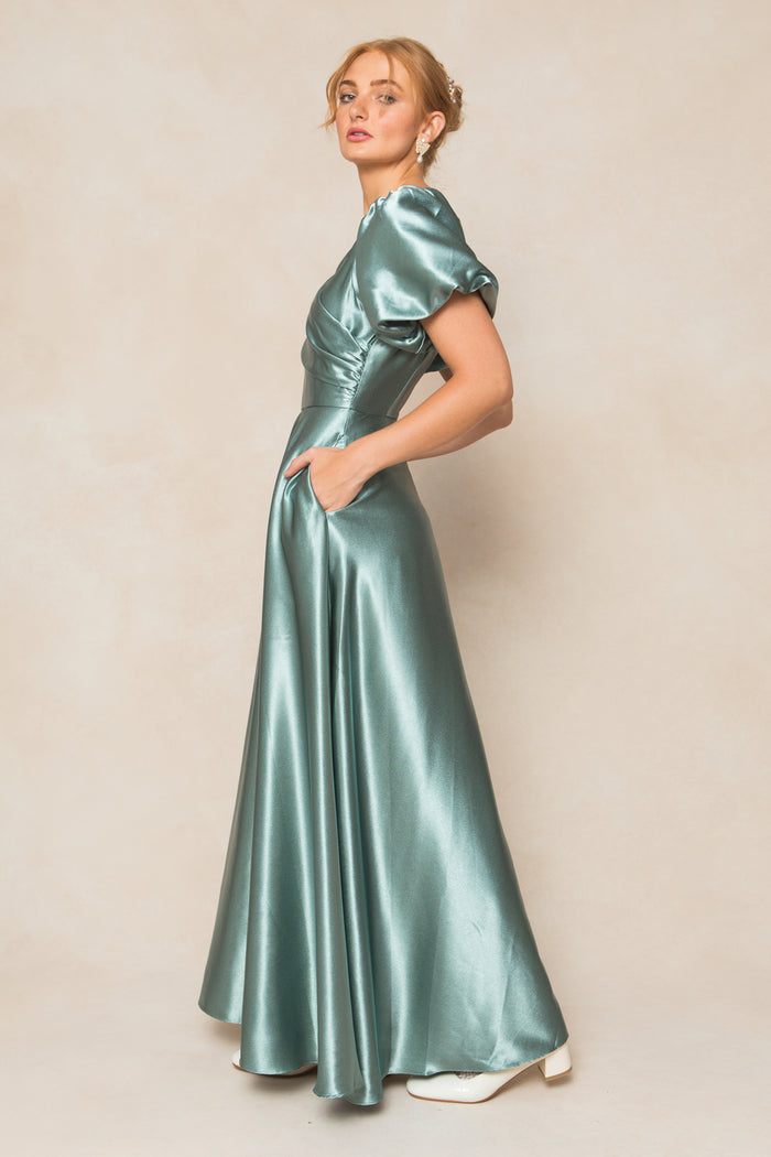 A woman in the Ivy City Co Tessie Dress in Seaglass Satin, featuring puff sleeves and pockets, stands sideways facing the camera. She pairs it with white shoes, statement earrings, and an updo against a neutral beige background.