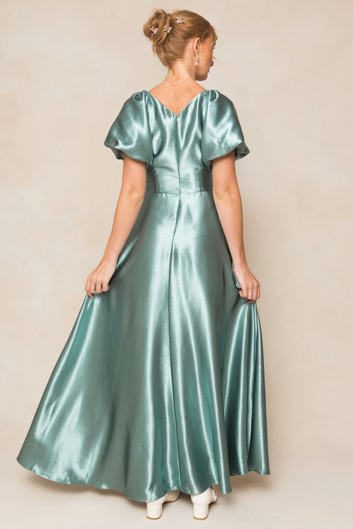 A woman with light hair in a bun, wearing the Ivy City Co Tessie Dress in Seaglass Satin—floor-length, short puff sleeves—stands with her back to the camera, holding out the skirt and wearing white heeled shoes; ideal for fall bridesmaids.
